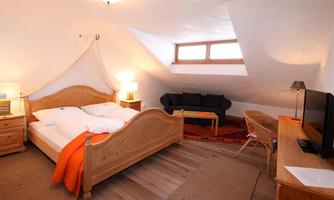 A cozy bedroom with a double bed and a small seating area. The walls are light and there are windows that let in natural light.