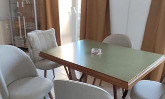 A bright dining area with a green table and several chairs. In the background, curtains and a television can be seen.