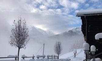 A wintry mountain landscape in the mountains with snow and fog. In the foreground, there are snow-covered trees and a wooden porch.