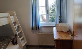 A bright children's room with a bunk bed and a wooden table. Light curtains hang in front of the window, creating a cozy atmosphere.