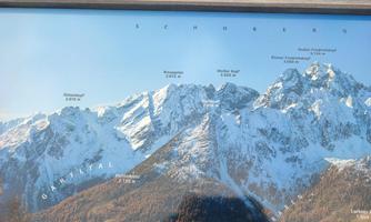 An impressive mountain panorama with snow-capped peaks and clear blue skies. In the foreground, the mountain names and elevation details are visible.