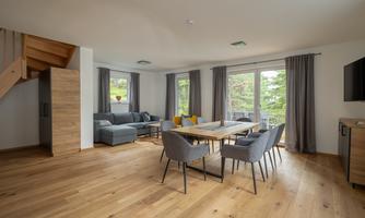 A modern living room with a comfortable couch and a dining area. Large windows provide plenty of natural light and a view of nature.