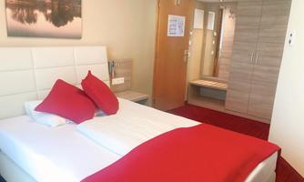 A cozy bedroom with a white bed and red pillows. In the background, there is a wardrobe and a picture of a landscape.