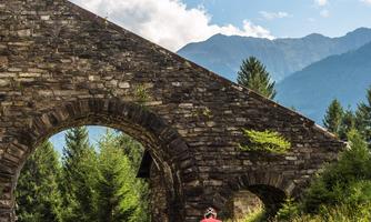 A picturesque cycling route leads through green meadows and under an old stone arch. In the background, mountains and a blue sky can be seen.