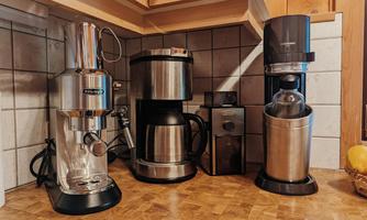 A kitchen unit with three different coffee machines. They are neatly arranged on a wooden shelf.