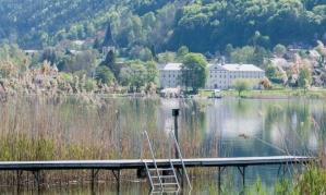 A tranquil lake with a pier in the foreground. In the background, green hills and a charming village can be seen.