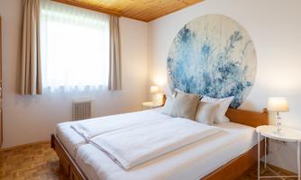 A cozy bedroom with a large double bed and a round wall painting. Daylight streams through the window, creating a bright atmosphere.