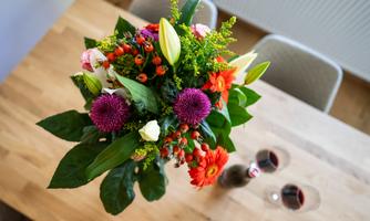 A colorful bouquet of flowers is on a wooden table. Two glasses of red wine are placed beside it.