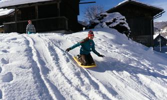 A person rides a sled down a snowy slope. In the background, there are wooden cabins and snow-covered landscapes.