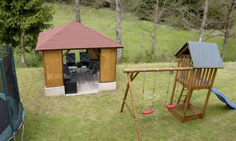 An outdoor playground with a play tower and a slide, as well as swings. In the background stands a cozy pavilion with seating.
