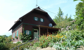 A rustic, red wooden house surrounded by blooming flowers and green plants. The building has green shutters and is located in an idyllic landscape.