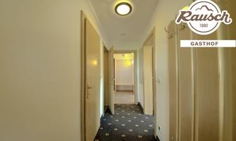 A bright hallway with closed doors and a blue carpet. At the end of the hallway, another door is visible that leads to another room.
