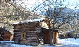 A rustic wooden house stands in a winter landscape. In the background, snow-covered trees and gentle hills are visible.