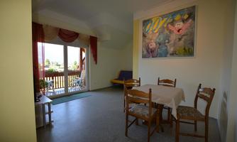 A bright room with a table and chairs as well as a colorful painting on the wall. Large windows offer a view of the balcony and the surroundings.