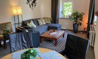 A cozy living room with a gray sofa, a wooden table, and large windows. Plants and artworks give the space an inviting atmosphere.