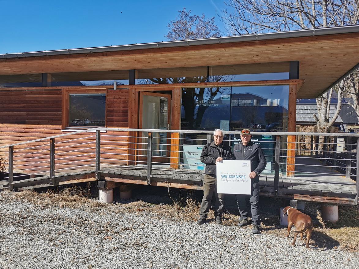 Weißensee zwei Männer halten Schild vor Holzhaus