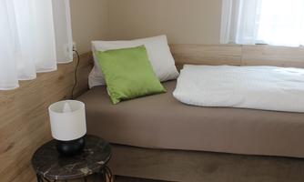 A cozy bedroom with a single bed and a green pillow. Next to the bed is a small table with a lamp.