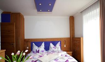 A modern bedroom with wooden furniture and colorful bedding. Large windows allow plenty of daylight in.