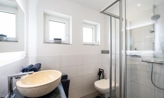 A modern bathroom with a large shower and a round sink. The walls are tiled in white and there are two small windows.