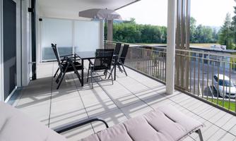A modern balcony with a table and chairs. In the background, there are trees and a quiet environment visible.