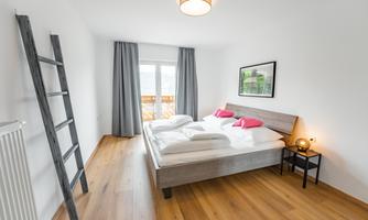 A cozy bedroom with a double bed and two pink pillows. Large windows let in plenty of daylight and the wooden floor creates a warm atmosphere.