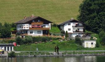 Two beautiful houses by the shore of a lake, surrounded by green landscape. On the balconies are chairs and tables that invite to a relaxed stay.