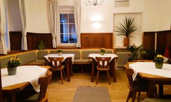 A cozy dining hall with bright tables and plants. The walls are made of wood, and a large chandelier illuminates the space.