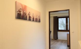 A simple hallway with a piece of art depicting birds on branches. Through the open door, a bright room with a window and curtains is visible.