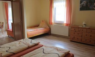 A bright and cheerful room with two beds and a single bed. The room has large windows with curtains and wooden furniture.
