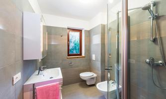 A modern bathroom with gray tiles and a shower. There is a sink, a toilet, and a window with a view to the outside.