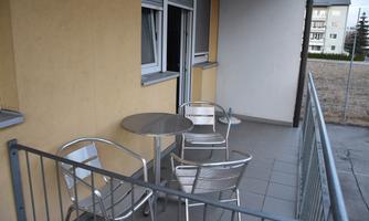 A small balcony with a round table and four metal chairs. The wall is painted in a light yellow, and a window opening is visible.