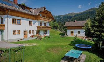 A beautiful alpine setting with two charming houses. The garden features a lawn area, a trampoline, and a table tennis court, surrounded by mountains.