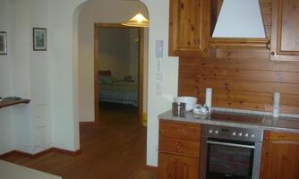 A cozy kitchen with wooden shelves and a stove. In the background, you can see an adjacent room with a bed.