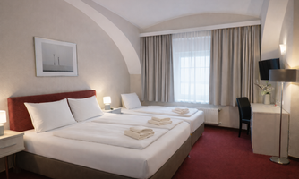 A hotel room with a double bed and a single bed, as well as a large window. The decor is simple and elegant, featuring carpet flooring and a desk.