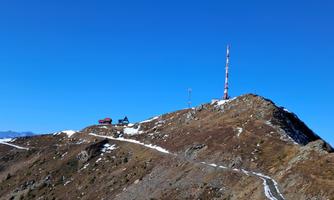 A mountain peak with a small cabin and a tall transmission tower. The sky is clear and blue, and remnants of snow are visible on the ground.