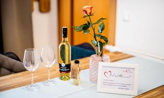 A set table with two wine glasses, a bottle of white wine, and a decorative vase with a rose. On the table lies a welcome sign that kindly greets.