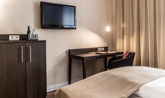 A modern hotel room with a bed, a television on the wall, and a desk. Next to the desk is a comfortable chair and there are curtains that let light in gently.
