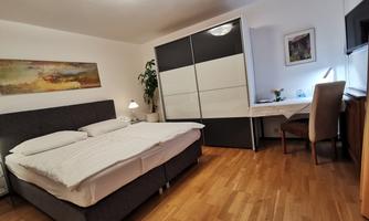 A cozy bedroom with a large bed and a desk. The room has a modern style and warm wooden flooring.