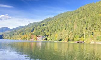 A tranquil lake surrounded by green hills and colorful trees in autumn. The sky is clear and blue, creating a peaceful atmosphere.