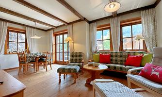 A cozy living room with a wooden ceiling and large windows. There is a seating area with a table, chairs, and comfortable sofas.