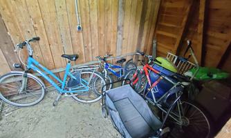 A small room with several bicycles in different colors. The floor is covered with dirt, and the walls are made of wood.