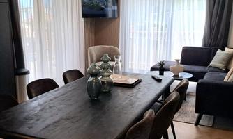 A modern living room with a large dining table and elegant chairs. The room is brightly lit by large windows and decorated with stylish furniture.