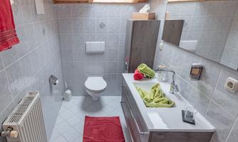 A modern bathroom with gray tiles and a wooden ceiling. There is a sink with towels in red and green, as well as a toilet.