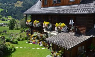 An idyllic wooden house with blooming flower balconies in a green landscape. The surroundings are characterized by gentle hills and trees.