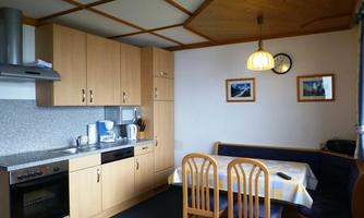A modern kitchen with wooden cabinets and a dining area. In the background, pictures are visible on the wall and kitchen appliances.