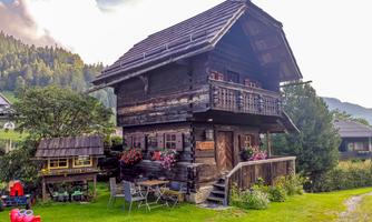 A traditional wooden house with a balcony, surrounded by green grass and trees. In the foreground are garden furniture and a toy vehicle.
