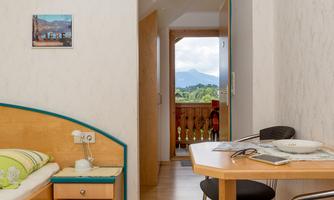A simple room with a single bed and a table. Through the door, there is a view of the mountains.