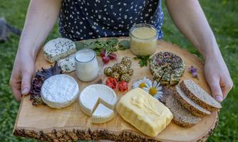 A rustic wooden board with various types of cheese, bread, and a small amount of yogurt. Surrounded by fresh herbs and edible flowers.
