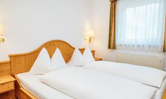 A cozy bedroom with a double bed and two upright pillows. The room has light walls and a large window with curtains.
