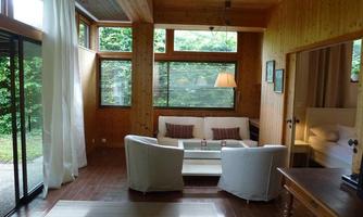 A cozy living room with wooden walls and large windows. The furnishings consist of a sofa, two armchairs, and a table, surrounded by natural light.